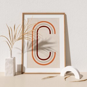May include: A framed print with a stylized letter "C" formed by overlapping orange, red, and gray arcs on a beige background. The print is in a wooden frame and is sitting on a light beige surface.