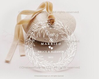 Könnte beinhalten: Ein transparenter Weihnachtsbaumschmuck aus Acryl mit einem weißen Kranzdesign und dem Text "Merry & Married" mit den Namen "Bruce & Eleanor" und dem Jahr "2022".