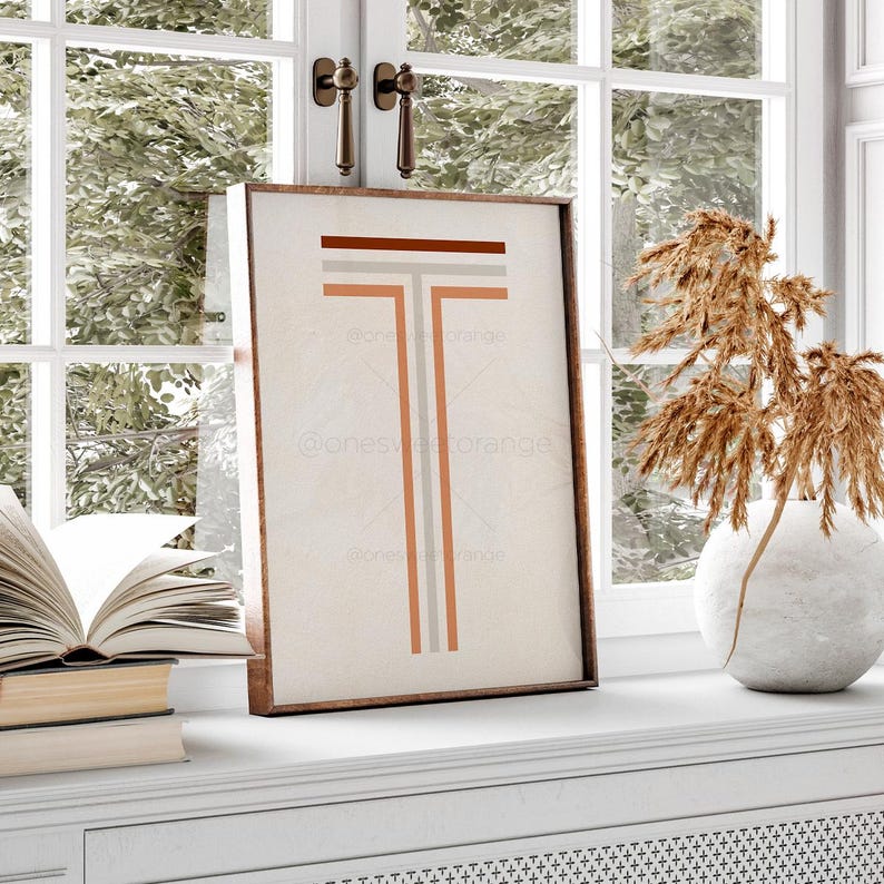 May include: A framed print with a geometric design of a letter T in shades of brown, orange, and gray on a cream background.