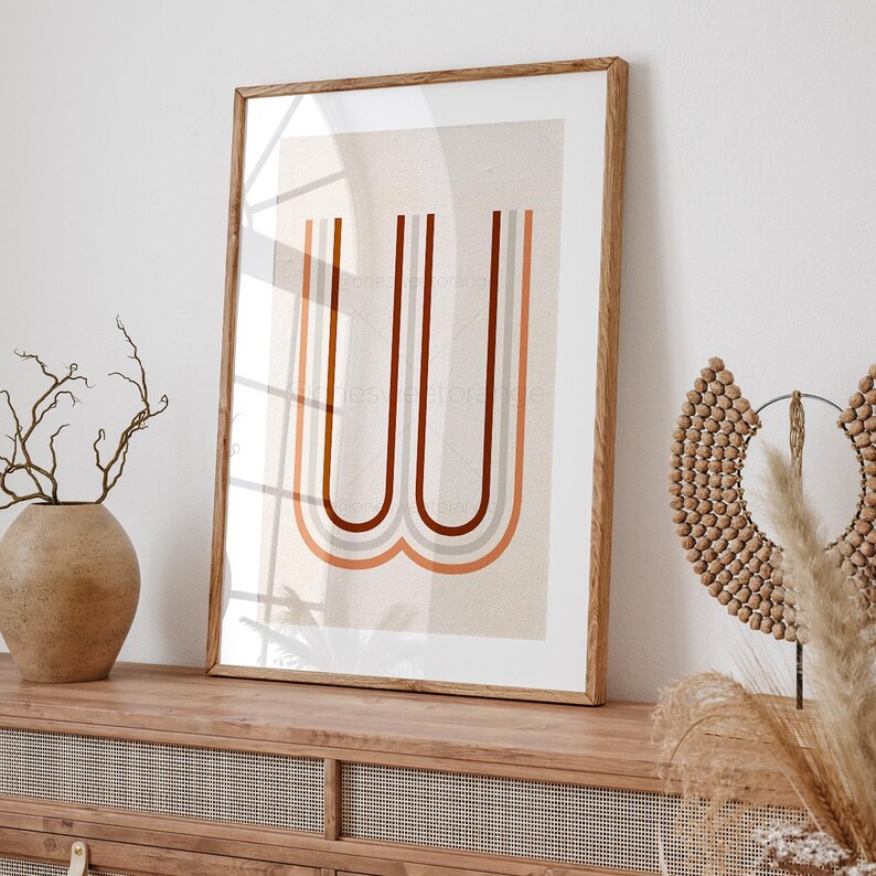 May include: A framed print with a minimalist design featuring a stylized letter "W" in a rainbow color palette of orange, red, brown, and gray. The print is on a beige background and is framed in a natural wood frame.
