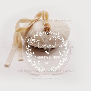 May include: Clear round ornament with a white floral wreath design and the text "Engaged" with two small hearts. Below the text is "Andrew & Jess" and the year "2021".