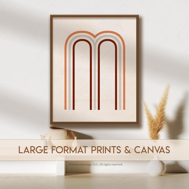 May include: A framed print of a minimalist abstract design featuring two arches in shades of brown, orange, and gray. The print is displayed on a white wall with a vase of dried pampas grass and a ceramic pot.