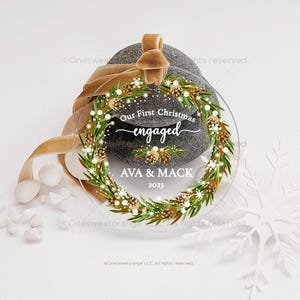 May include: A clear round Christmas ornament with a green pine wreath and white snowflakes. The ornament has the text "Our First Christmas engaged AVA & MACK 2023" on it.