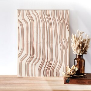 May include: A framed print with a brown and white abstract pattern of wavy lines. The print is sitting on a wooden surface with a vase of dried pampas grass and a book titled "Club House" in front of it.