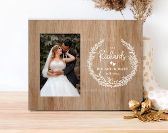 May include: A wooden picture frame with a wedding photo of a couple in a forest setting. The frame has a white inscription that reads "The Richards, Roland & Mary, 9.18.2024" with a wreath design.