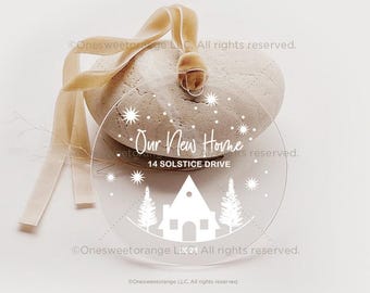 May include: A clear round ornament with a white house, trees, and snowflakes. The text "Our New Home" and "14 Solstice Drive" is printed on the ornament.