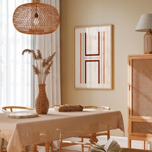 May include: A light brown wicker pendant lamp hangs over a dining table with a beige tablecloth. A framed print with a geometric design in shades of red, orange, and gray hangs on the wall behind the table. A wicker cabinet with a light wood frame is visible in the background.