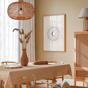 May include: A light brown wicker pendant lamp hangs over a dining table with a beige linen tablecloth. A framed print with a black and white sun and moon design hangs on the wall behind the table. A wicker cabinet with a light brown wood frame is in the background.