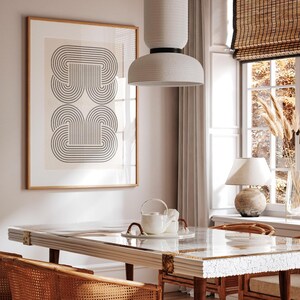 May include: A white ceramic pendant light with a geometric design hangs over a dining table with a glass top and a white marble base. The table is set with a white teapot and two white cups. The room has a light beige wall with a framed print of a black and white geometric design.