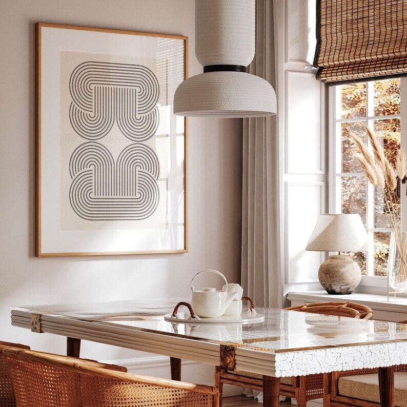 May include: A white ceramic pendant light with a geometric design hangs over a dining table with a glass top and a white marble base. The table is set with a white teapot and two white cups. The room has a light beige wall with a framed print of a black and white geometric design.