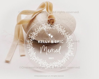 Könnte beinhalten: Ein runder, transparenter Anhänger mit einem weißen Kranz-Design und dem Text "KELLY & ERIC Married 2021" im Inneren. Der Anhänger hängt an einem goldenen Band.