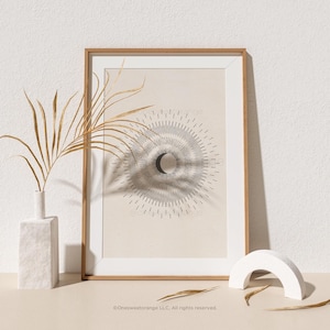 May include: A framed print with a beige background and a black and white illustration of a crescent moon surrounded by radiating lines. The print is in a light wood frame and is sitting on a table with a white vase and a white arch.