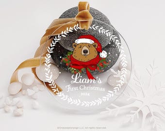 May include: Clear round Christmas ornament with a white wreath border. Inside the wreath is a cartoon illustration of a brown bear wearing a red Santa hat and scarf. The text "Liam's First Christmas 2024" is printed below the bear.