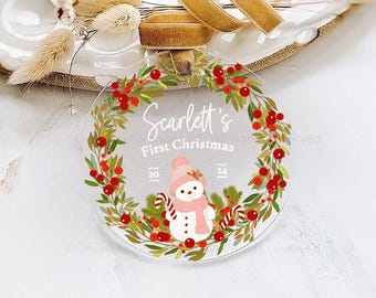 May include: A clear round ornament with a red and green wreath design. The wreath has red berries and green leaves. In the center of the wreath is a snowman with a pink hat and scarf. The text "Scarlett's First Christmas" is written above the snowman. The years 20 and 24 are written on either side of the text.