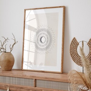 May include: A framed print with a minimalist design of a crescent moon surrounded by radiating lines. The print is on a light beige background and is framed in a natural wood frame. The print is displayed on a wooden shelf with a decorative wooden piece and dried grasses.