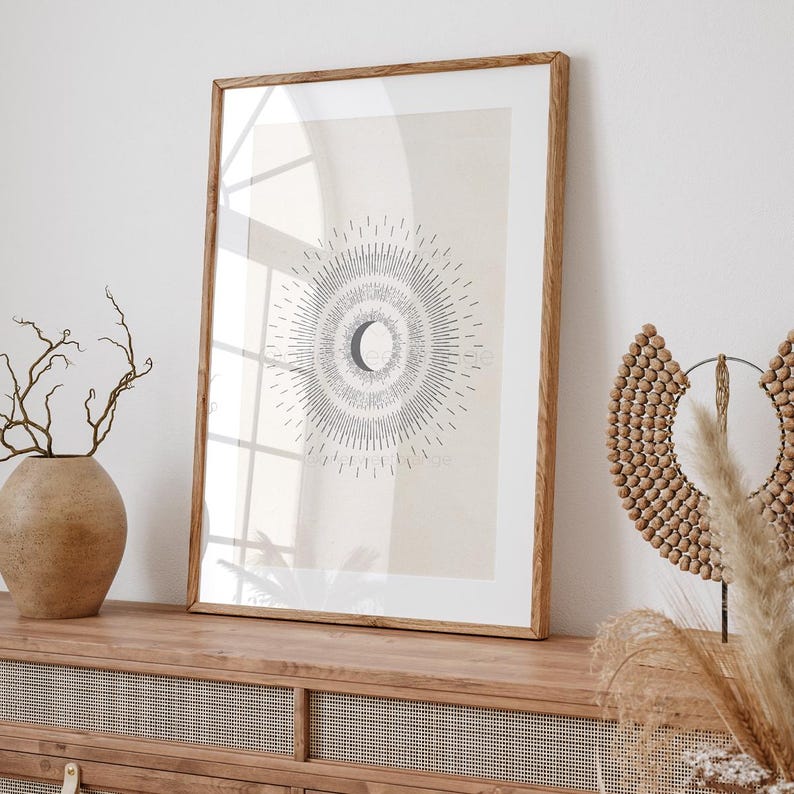May include: A framed print with a minimalist design of a crescent moon surrounded by radiating lines. The print is on a light beige background and is framed in a natural wood frame. The print is displayed on a wooden shelf with a decorative wooden piece and dried grasses.