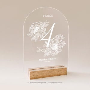 May include: White acrylic table number with floral design and the text "TABLE 4 Madelyn & Robert 10.10.2026" on a wooden base.