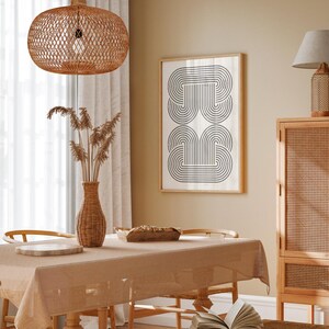 May include: A dining room with a light brown table and chairs, a woven pendant light, a vase with dried flowers, and a framed black and white geometric print on the wall.