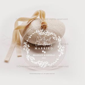 May include: Clear round ornament with a white wreath design and the text "Our First Christmas Married" with the names Bradley & Kendal and the year 2021.