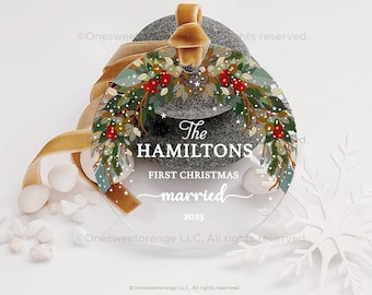 May include: A clear round Christmas ornament with a green and red floral wreath design. The text "The Hamiltons First Christmas married 2023" is printed on the ornament.