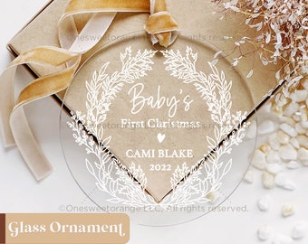 Puede incluir: Un adorno de vidrio transparente con un diseño de corona de flores blancas. El adorno dice "Baby's First Christmas" con un corazón y el nombre "CAMI BLAKE" y el año "2022".