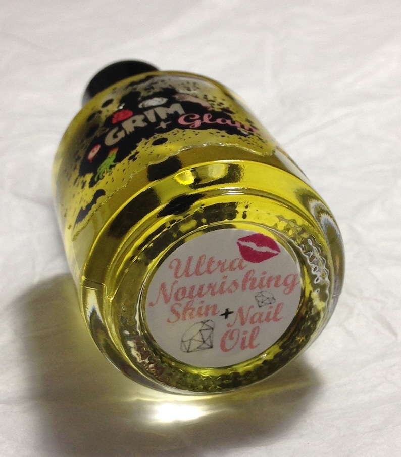 Op de afbeelding: Een heldere glazen fles gevuld met gele vloeistof, met het label "Grim Glam" met een zwarte dop. Het onderste label luidt "Ultra Nourishing Skin + Nail Oil" met een rode lip-afbeelding en een diamantillustratie.