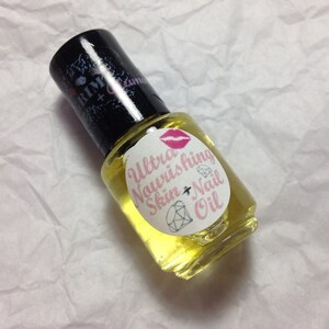 Op de afbeelding: Een kleine glazen fles Ultra Nourishing Skin + Nail Oil met een zwarte dop. Het etiket heeft roze tekst, een diamant en lippen. De olie is lichtgeel van kleur.