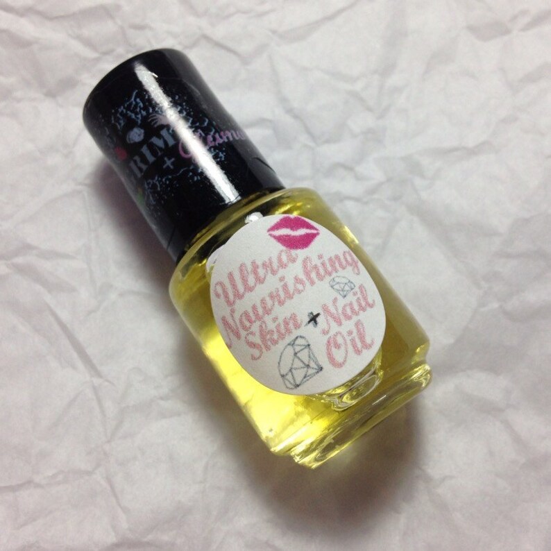 Op de afbeelding: Een kleine glazen fles Ultra Nourishing Skin + Nail Oil met een zwarte dop. Het etiket heeft roze tekst, een diamant en lippen. De olie is lichtgeel van kleur.