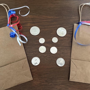 May include: Two rectangular brown paper gift bags with twisted paper handles, each decorated with curled red, white, and blue ribbons. Between the bags are six silver-colored coins of varying sizes arranged in a symmetrical pattern on the surface.