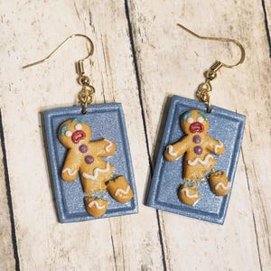 May include: A pair of gingerbread man earrings. Each earring features a light brown gingerbread man with white icing details, purple buttons, and a blue frame. The earrings have gold-colored hooks.