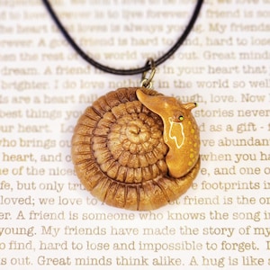 May include: A gold snail charm necklace with a brown and gold snail pendant. The snail is curled up with its shell facing the viewer. The necklace is on a black cord.