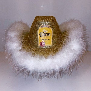 Può includere: Un cappello da cowboy in glitter dorato con un bordo di piume bianche. Il cappello ha un'etichetta Jose Cuervo Especial sul davanti.