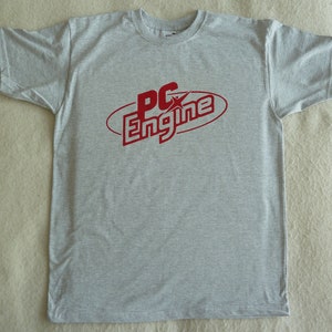 Puede incluir: Una camiseta gris con un logotipo rojo que dice "PC Engine" en una fuente estilizada.