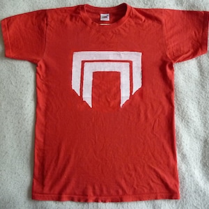 May include: Red t-shirt with a white graphic of a stylized, abstract design. The design is made up of three overlapping rectangles.