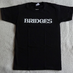 Può includere: T-shirt nero con la scritta "BRIDGES" stampata in bianco sul petto. La maglietta ha maniche corte e girocollo. Il tessuto sembra essere un morbido misto cotone.
