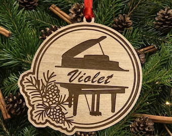 Pode incluir: Um enfeite de Natal de madeira em forma de piano de cauda com o nome "Violet" gravado nele. O enfeite é decorado com um ramo de pinheiro e pinhas.