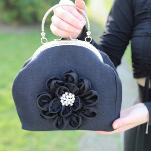 May include: A black fabric clutch bag with a silver clasp and a large black fabric flower with a rhinestone center.