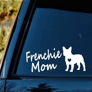 Pu&ograve; includere: Adesivo in vinile bianco con il testo "Frenchie Mom" e una silhouette di un Bulldog Francese con un cuore sul petto. L'adesivo &egrave; su un finestrino di un'auto blu.