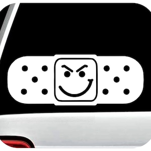May include: White vinyl sticker of a band-aid with a smiley face with an evil grin. The sticker is on a black car window.