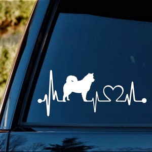 May include: White vinyl decal of a dog silhouette with a heartbeat line and a heart. The decal is on a blue car window.