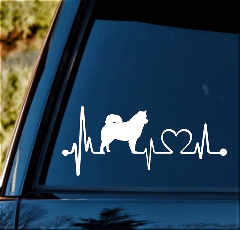 May include: White vinyl decal of a dog silhouette with a heartbeat line and a heart. The decal is on a blue car window.