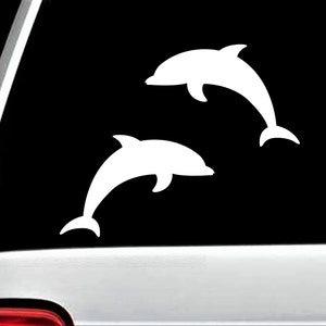 May include: Two white dolphin silhouettes are depicted against a black background. The dolphins are in a jumping pose, with their bodies arched and tails curved. The image is likely a decal or sticker.