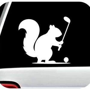 May include: White vinyl decal of a squirrel playing golf. The squirrel is holding a golf club and is positioned to hit a golf ball. The image is set against a black background.
