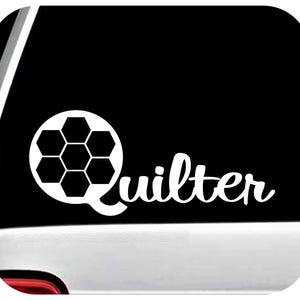 May include: White decal featuring a soccer ball design and the word "Quilter" in a cursive font. The decal is likely for a car window or similar surface, with a black background.