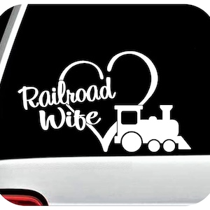 May include: White vinyl decal featuring the words "Railroad Wife" in a cursive font, integrated with a heart shape and a train illustration. The design is set against a black background.