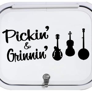 May include: White rectangular sign with the text "Pickin' & Grinnin'" in black script. Silhouettes of a guitar, violin, and banjo are to the right of the text. A metal latch is at the bottom.
