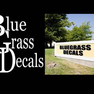 May include: A yellow sign with white trim and black text that reads "BLUEGRASS DECALS" in front of a grassy lawn.