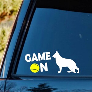 May include: White vinyl decal featuring the words "GAME ON" in bold white letters, a yellow tennis ball, and a white silhouette of a German Shepherd dog. The decal is designed for application on a car window.