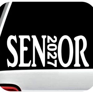 May include: White "SENIOR 2027" decal on a black background. The text is in a bold, stylized font, suitable for a car window or similar application. The decal is displayed on a white surface.
