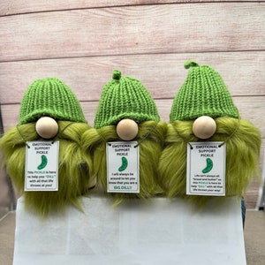 May include: Three green gnomes with long green beards and green knit hats. Each gnome has a sign hanging from its neck that says "Emotional Support Pickle" with a different pickle-themed message on each sign.